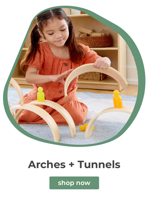Child playing with a wooden arch and tunnel toy on a carpeted floor.