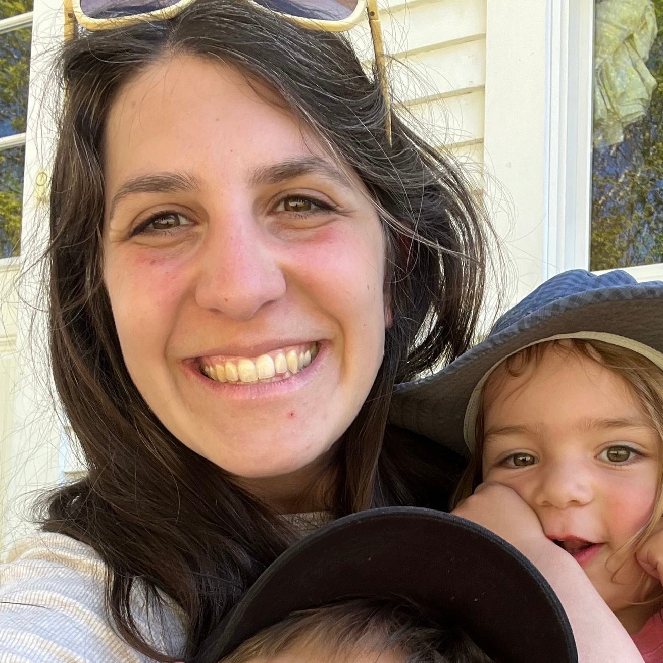 A smiling woman with two young children, one in a sun hat, poses closely together outdoors in warm sunlight.