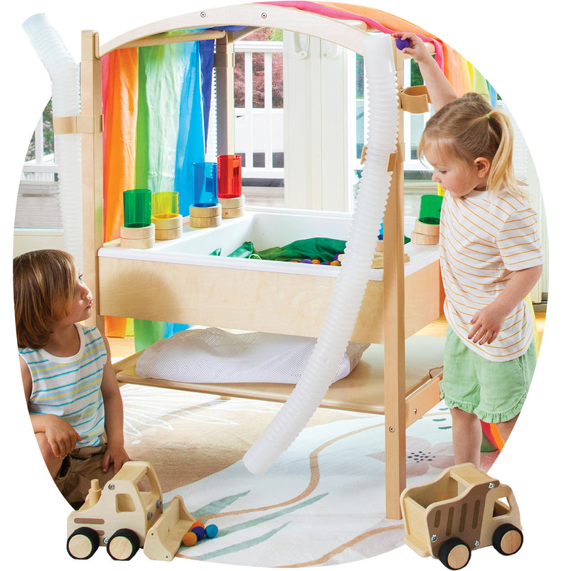 A children's wooden play structure with a white trough and colorful hanging fabric panels, shown in a bright indoor room with two children playing around it. The scene is cropped into a circular frame with toy trucks visible in front.