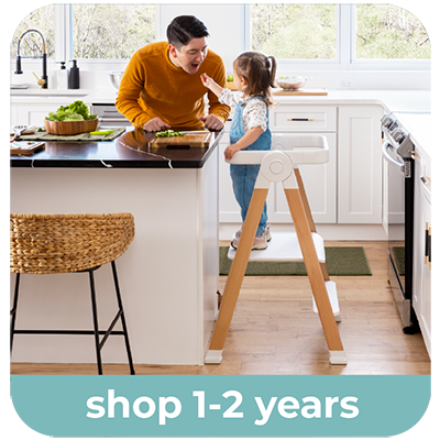 A toddler stands in a wooden and white Kitchen Helper Flex in a bright kitchen, reaching toward her father who is standing nearby. A light teal banner at the bottom reads 'shop 1-2 years'.
