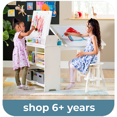 A children's white writing and drawing drafting desk set shown in a bright room with two young girls using it. One girl stands at the easel writing on a white board, and the other sits at the writing surface holding a red drawing pad. Shop 6+ in text below image