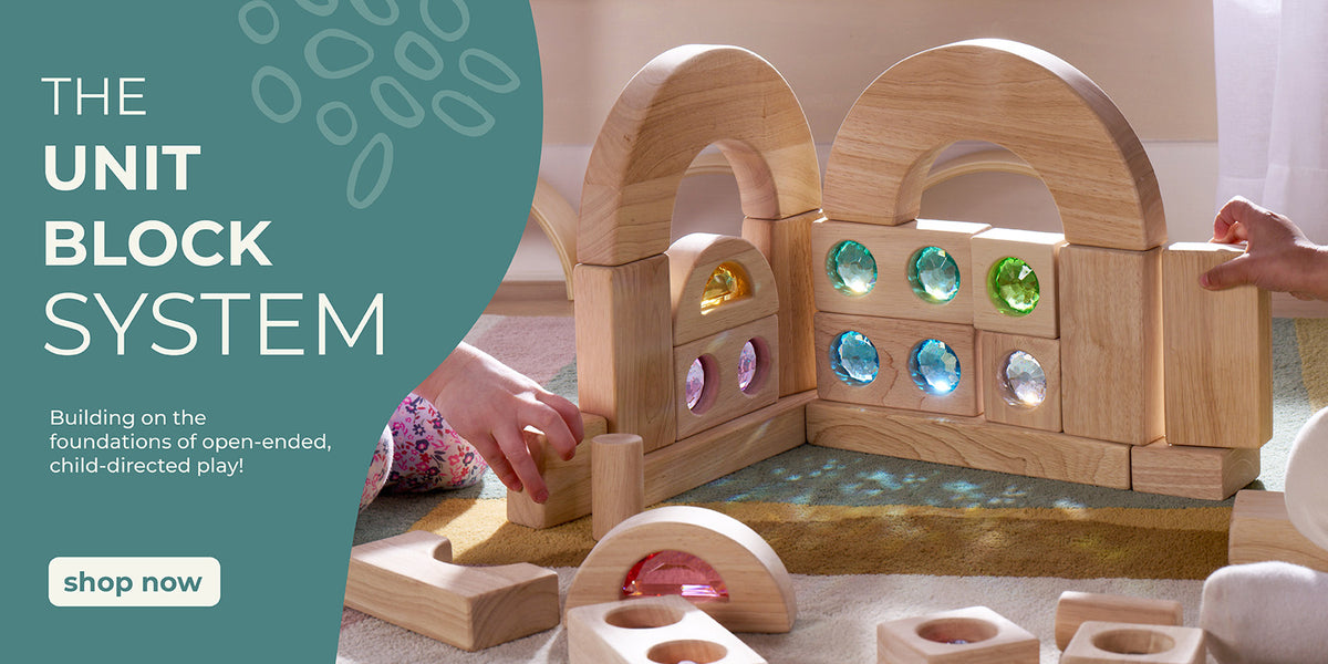Wooden unit block set with light shining through colorful, gem-like acrylic inserts and two children's hands visible placing them together to create a beautiful structure. Unit Block System promotional text on the left with a call to action button to shop the line.