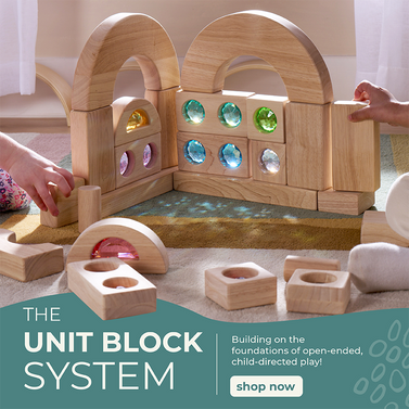 Wooden unit block set with light shining through colorful, gem-like acrylic inserts and two children's hands visible placing them together to create a beautiful structure. Unit Block System promotional text on the left with a call to action button to shop the line.