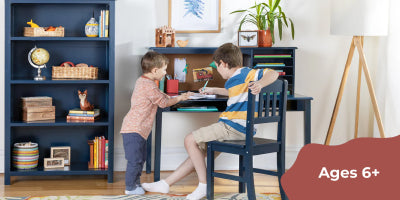 Two boys collaborate at a desk in a vibrant room with shelves; text reads Ages 6+, encouraging teamwork.