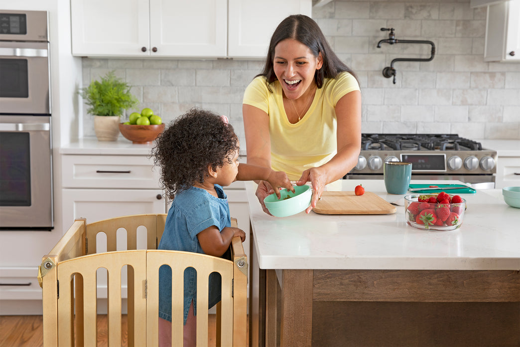 Mom giving daughter a bowl of fruit at a kitchen counter. Click to shop the Kitchen Helper toddler tower.