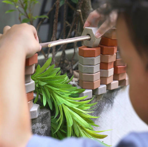 A person threads a stick through hexagonal ceramic pieces stacked on a wall near lush green plants.