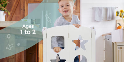 Smiling toddler stands behind a white baby gate in a bright kitchen, with text that reads Ages 1 to 2 and safety at home.