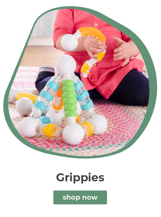A child wearing a pink shirt sits on a patterned rug indoors while playing with a colorful plastic toy structure labeled 'Grippies' and a green 'shop now' button below on a white background.