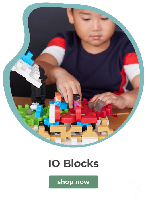 A child wearing a navy, red, and white shirt is focused on building with colorful toy blocks. The image is framed in a rounded teal shape with the text 'IO Blocks' and a green 'shop now' button below on a white background.